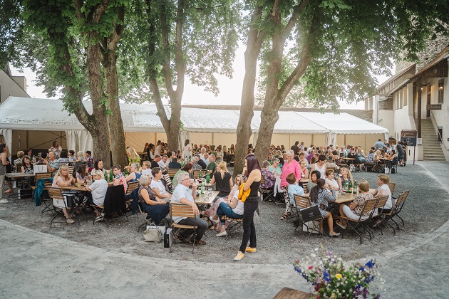 Ein Bild vom Sommerfest. Mitarbeitende sitzen an Tischen draußen im Freien unter den Bäumen.